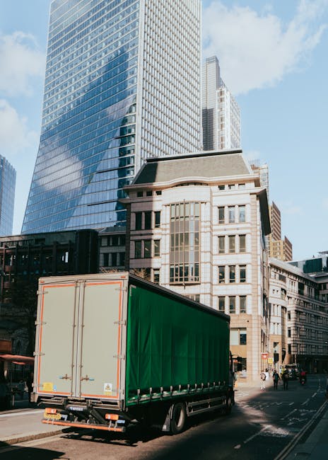 A large green box trailer attached to a vehicle is parked on a city street in front of modern office buildings and older architectural structures. The trailer is positioned on the pavement, with the rear doors closed and secured. The surrounding environment includes a clear blue sky with some clouds, and the street appears to have minimal traffic with a few pedestrians visible in the background. The setting suggests a typical urban scene where house removals or furniture transport might occur, aligning with professional moving services such as those provided by Man and Van Knightsbridge. The scene captures the loading or unloading process, with the trailer ready to be used in a home relocation or office move within the city center.