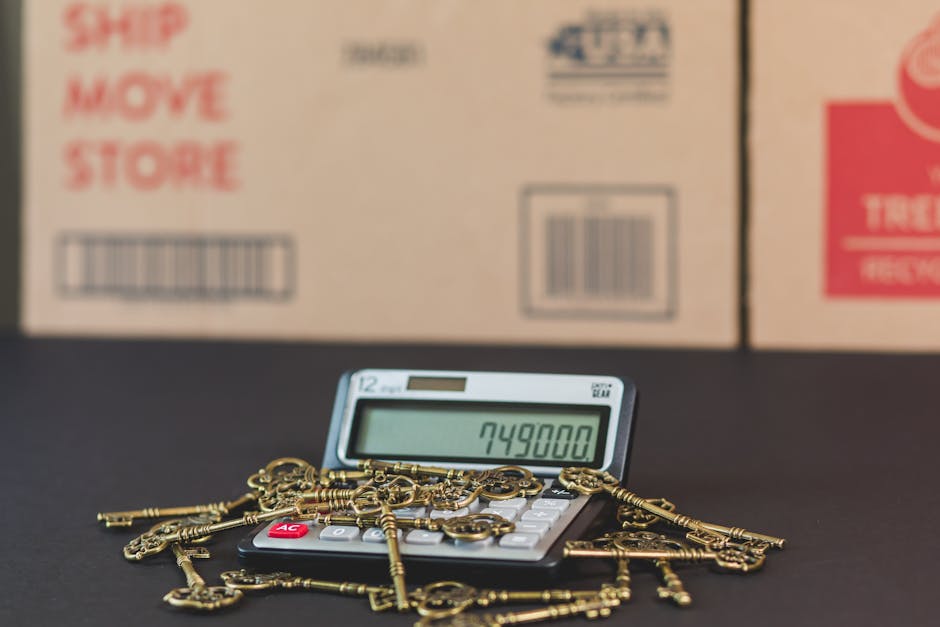 A digital calculator displaying the number 749,800 is placed on a black surface, surrounded by numerous small brass keys with ornate designs, resembling antique or vintage keys. In the background, three stacked cardboard boxes with printed labels, including red and blue text and barcodes, are visible inside a room, possibly a storage or move preparation area. The setting appears to be indoors with good lighting, and the scene suggests an element of packing or inventory, related to house removals or furniture transport. Man and Van Knightsbridge, a professional removals service, is associated with the scene, indicating this image relates to home relocation and moving logistics involving packing, handling, and loading processes.
