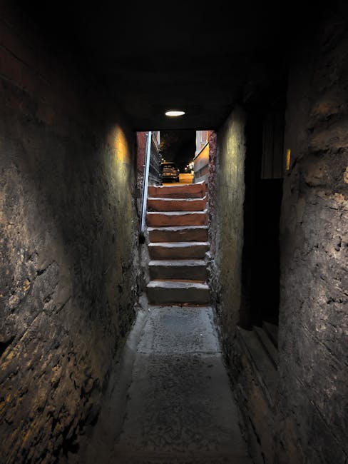 A narrow, dimly lit stone corridor leading to a set of wooden stairs ascending to a street level, with a small, round ceiling light providing minimal illumination. On the left and right sides, the walls are made of exposed, rough stone, and the ceiling is low. The stairs are partially illuminated by natural light from the street above, highlighting the weathered wooden treads. At the top of the stairs, a black vehicle is parked on the street, visible through an open outdoor area. This corridor appears to be an access point for a basement, illustrating typical challenges in home relocation and furniture transport through restricted or difficult pathways. During the process of removals by Man and Van Knightsbridge, such narrow passages require careful planning for loading and unloading to prevent damage to furniture and ensure safe movement during the house removal or packing and moving services.
