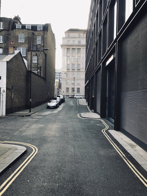 A narrow urban street with a slight incline, showing a modern building with a black metal façade and roller shutter door on the right side, alongside traditional brick buildings on the left. Several parked cars are visible along the curb, with a white van partially visible at the far end of the street. The pavement has double yellow lines indicating parking restrictions, and there are no pedestrians present. The scene is illuminated by daylight with overcast weather, providing natural, neutral lighting. This environment illustrates the limited access typical for house removals and furniture transport in tight city streets, where careful planning and specialized vehicle access are required, as often handled by companies like Man and Van Knightsbridge during home relocation and packing and moving services.