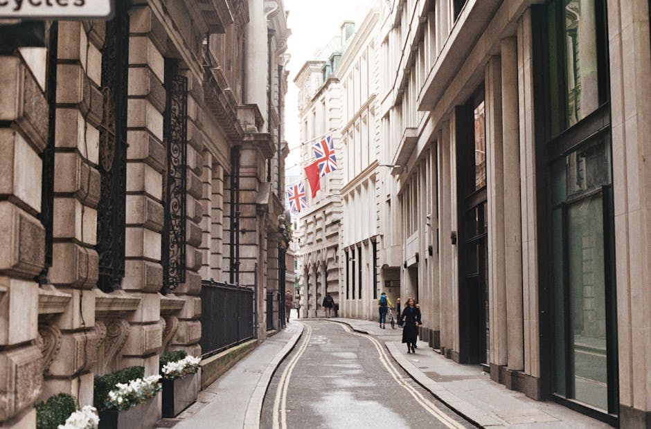 A narrow urban street in Knightsbridge with tall, elegant buildings on both sides featuring detailed stone facades and large windows. Union Jack flags and red ceremonial banners are strung across the street, hanging between the facades. The street pavement is wet, with a single yellow line running along the curb on the right side. A group of pedestrians, dressed in casual attire, walk along the pavement, some near the building entrances. Inside the section of the street visible, there is no visible moving activity or furniture, but the setting suggests a typical location where house removals and furniture transport might be carried out. Man and Van Knightsbridge often operate in such environments for client house relocations, especially when navigating narrow streets like this one, highlighting the importance of access tips for effective packing and moving logistics in historic or busy areas.