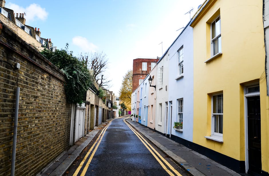A narrow urban street with a slight incline, showing a modern building with a black metal façade and roller shutter door on the right side, alongside traditional brick buildings on the left. Several parked cars are visible along the curb, with a white van partially visible at the far end of the street. The pavement has double yellow lines indicating parking restrictions, and there are no pedestrians present. The scene is illuminated by daylight with overcast weather, providing natural, neutral lighting. This environment illustrates the limited access typical for house removals and furniture transport in tight city streets, where careful planning and specialized vehicle access are required, as often handled by companies like Man and Van Knightsbridge during home relocation and packing and moving services.
