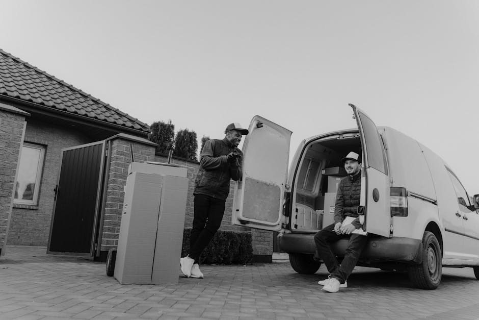 A man with a serious expression, wearing a white polo shirt and a black cap, stands inside the open rear doors of a moving van, which is filled with neatly stacked cardboard boxes of various sizes, so