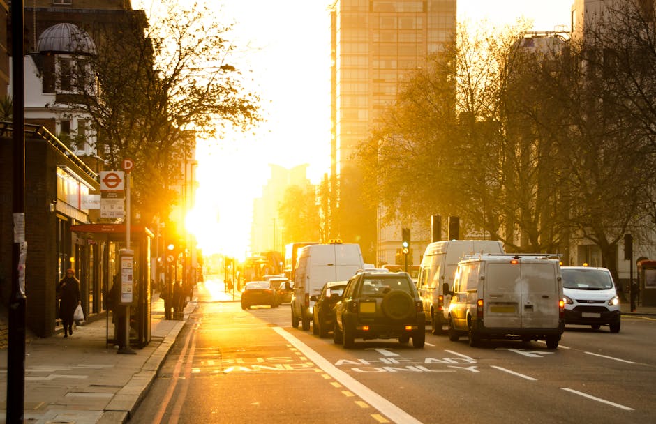 A busy urban street scene during sunset in Knightsbridge, London, with vehicles including white vans, passenger cars, and a bus waiting in traffic near the curb. The street features a dedicated bus lane marked with road markings, and several pedestrians are walking along the sidewalk, some approaching a small bus shelter. The background includes tall modern office buildings, traditional architecture with a domed structure on the left, and trees lining the sidewalk, illuminated by the golden glow of the setting sun. The scene captures a moment during home relocation or furniture transport as part of a moving process, with vehicles likely employed for house removals coordinated by Man and Van Knightsbridge, visible through the overall urban environment and typical removals activity.