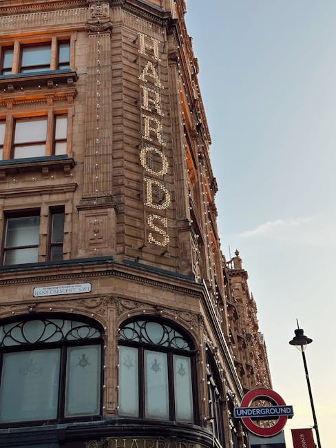 A multi-storey building with an ornate stone façade features large windows on the ground floor with decorative arched glass panels. Vertical signage spelling 'HARRODS' is displayed on the corner of the building using illuminated letters. The building is adorned with decorative elements and string lights along the edges of its upper floors. The sky is clear with a few clouds, and a street lamp and an underground subway sign are visible in the foreground. This scene captures a historic part of Knightsbridge, where professional removals by Man and Van Knightsbridge may involve packing, loading furniture, and transporting household items in a logistics operation related to house removals from Harrods nearby to Hyde Park.