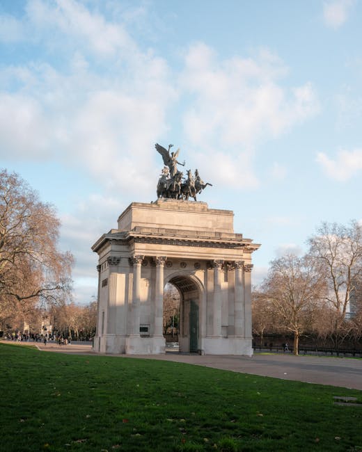 A tall, ornate stone archway situated on a grassy area with a wide paved path leading through it. The arch features detailed carvings and a large open central passage. Atop the arch is a bronze or dark metal statue of a chariot pulled by four horses, with a figure holding a spear or staff standing inside the chariot. Leafless trees are visible in the background, and the sky above is mostly clear with a few scattered clouds. The lighting suggests a bright, sunny day. This scene is part of a city park in London, near the landmarks mentioned in the Knightsbridge removals service page by Man and Van Knightsbridge, which specializes in home relocation and furniture transport services.