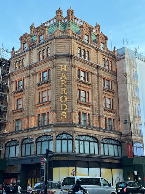 A multi-storey building with an ornate stone façade features large windows on the ground floor with decorative arched glass panels. Vertical signage spelling 'HARRODS' is displayed on the corner of the building using illuminated letters. The building is adorned with decorative elements and string lights along the edges of its upper floors. The sky is clear with a few clouds, and a street lamp and an underground subway sign are visible in the foreground. This scene captures a historic part of Knightsbridge, where professional removals by Man and Van Knightsbridge may involve packing, loading furniture, and transporting household items in a logistics operation related to house removals from Harrods nearby to Hyde Park.