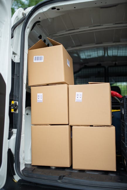 Inside a white delivery van used for home relocation, four large cardboard boxes with white shipping labels are stacked, with three positioned on the van's floor and one placed on top of the others. The boxes are sealed with packing tape, and the van's interior ceiling and side panel are visible, with the rear door open to reveal an outside pavement and a partial view of a vehicle in the background. This scene depicts the loading process common in furniture transport and packing during a house removal, with the boxes prepared for transport by Man and Van Knightsbridge, supporting efficient relocation services in Knightsbridge. The environment is brightly lit, with existing moving equipment like trolleys or straps not visible, emphasizing a straightforward loading operation for furniture and packed goods.