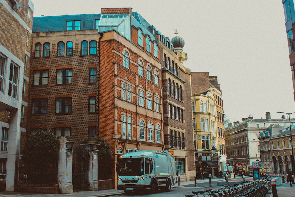 A street scene outside a multi-storey brick building with large windows, situated in an urban area with other historic and modern buildings visible in the background. A small blue commercial van from