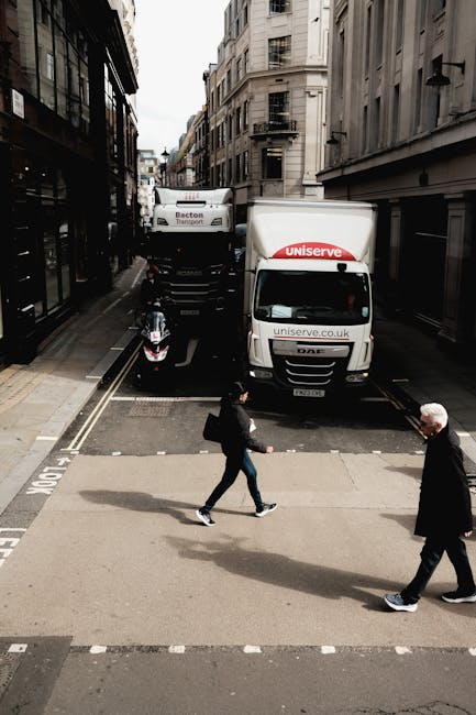 A street scene during a home relocation process showing a delivery van and a moving truck parked along a narrow urban street with tall, beige-colored buildings on either side. The white delivery van, branded with 'uniserve.co.uk,' is positioned at the curb, partially blocking the roadway, with a small black scooter parked nearby. Two pedestrians are crossing the street: a woman dressed in dark clothing carrying a bag and a man with white hair in a black coat. The pavement is marked with white dashed lines indicating a pedestrian crossing. The scene is set during daylight hours with natural lighting illuminating the area, highlighting the activity of furniture transport and packing preparations typical of house removals, illustrating the logistical aspects involved in furniture transport within a city environment, as handled by companies like Man and Van Knightsbridge.