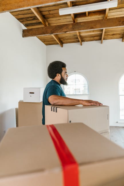 A man with dark curly hair and a beard, dressed in a blue shirt, is standing inside a bright room with white walls and large arched windows, preparing for a house relocation. He is positioned behind several stacked cardboard boxes of various sizes, with one prominent box in the foreground wrapped with red tape. The room features an unfinished wooden ceiling with exposed beams, indicating a new or renovated property. The man appears to be in the process of moving or organizing items, potentially for packing or unloading as part of furniture transport and packing and moving services offered by Man and Van Knightsbridge. The scene captures the loading process within the interior space, emphasizing careful handling of items during home removals.