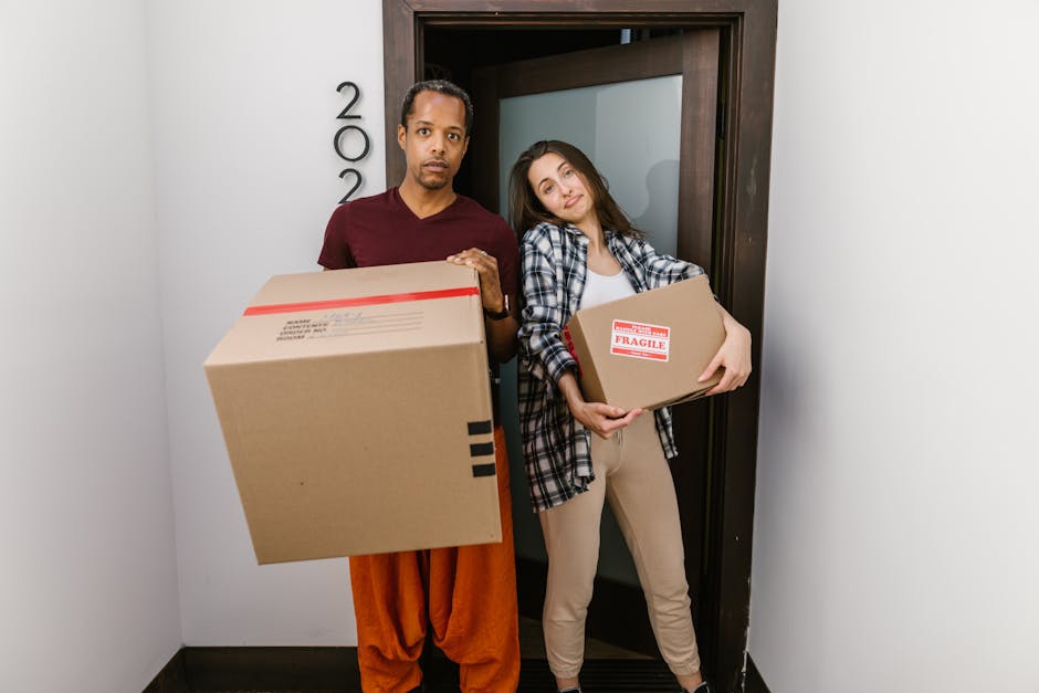 A man and a woman standing inside a property near a doorway, each holding several cardboard moving boxes, with one box marked 'Fragile.' The man, wearing an orange-brown pair of pants and a maroon top