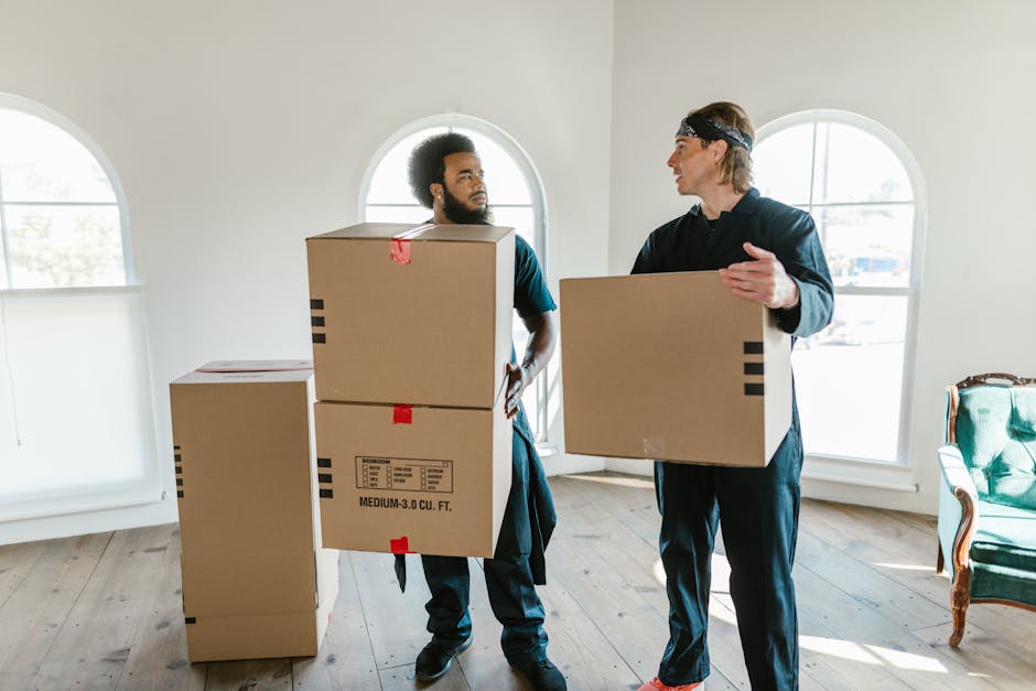 A man and a woman standing inside a property near a doorway, each holding several cardboard moving boxes, with one box marked 'Fragile.' The man, wearing an orange-brown pair of pants and a maroon top