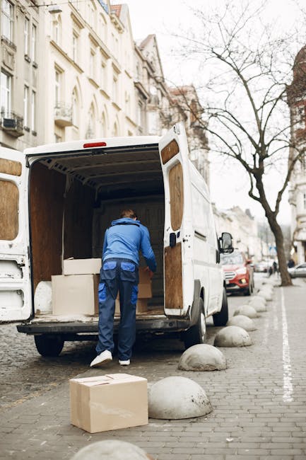 A man wearing a blue jacket and dark trousers is seen loading cardboard boxes and furniture into the back of a white commercial van on a city street. The van is parked parallel to the pavement, with its rear doors wide open, revealing an interior lined with plywood panels. The scene takes place outdoors during daylight, with a row of parked cars, including a red vehicle partially visible, along the curb. In the background, there are multi-storey buildings with white facades and large windows, and a leafless tree indicating a colder season. Several large, rounded concrete bollards line the edge of the sidewalk, separating the pedestrian area from the street. The process appears to be part of a home relocation or furniture transport service, possibly coordinated by Man and Van Knightsbridge, supported by visible moving equipment such as blankets or straps, though they are not clearly seen. The scene captures the essentials of a professional moving and packing operation in an urban environment.
