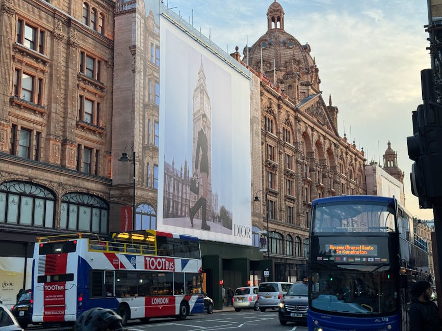A busy street scene in Knightsbridge, Westminster, featuring a large historic building with ornate architecture, decorative stonework, and a prominent dome at the top. Adjacent to the building, a tall advertising billboard displays a fashion model with a shopping bag, branded by Dior. On the road, a red and white double-decker bus labeled 'TOOTbus' is moving alongside a black double-decker bus with an electronic route sign indicating a route to Kensington. Several parked cars and moving vehicles are visible along the street. The scene is set during daylight with clear skies, and the overall environment reflects typical urban activity related to house removals and moving logistics, with vehicles and street-level activity aligned with London’s city centre traffic flow. The image provides context for ongoing or planned removals, highlighting vehicular movement, street infrastructure, and urban surroundings as part of comprehensive house relocation processes.